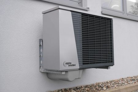 An Alpha Innotec heat pump unit mounted on a gray exterior wall above a gravel strip.