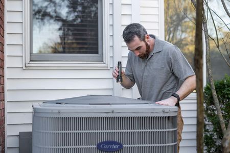 A technician uses a flashlight to inspect a Carrier air conditioning unit outside a residential house.