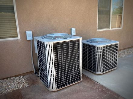 Two light-colored, boxy air conditioning units sit outdoors against a beige exterior wall near two windows.