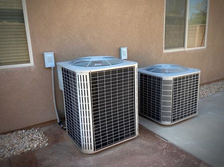 Two light-colored, boxy air conditioning units sit outdoors against a beige exterior wall near two windows.