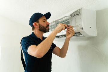 A technician in a blue uniform uses a screwdriver to service a white wall-mounted air conditioning unit.